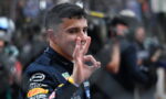 Race car driver in a dark suit grins and makes an OK sign with his right hand, crowd blurred in the background.