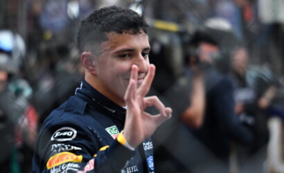 Race car driver in a dark suit grins and makes an OK sign with his right hand, crowd blurred in the background.