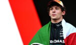 Young race car driver wearing a Pirelli cap, with the Italian flag draped over his shoulders and a black racing suit with sponsor logos.