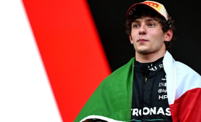 Young race car driver wearing a Pirelli cap, with the Italian flag draped over his shoulders and a black racing suit with sponsor logos.