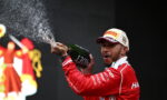 Racing driver on a podium spraying champagne from a bottle, wearing a red suit and a white Pirelli cap.