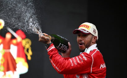 Racing driver on a podium spraying champagne from a bottle, wearing a red suit and a white Pirelli cap.
