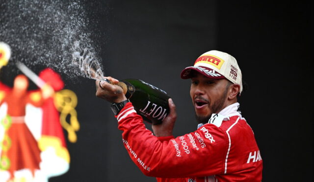 Racing driver on a podium spraying champagne from a bottle, wearing a red suit and a white Pirelli cap.