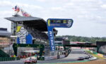 Race cars on a track with a large Michelin arch and blue signage near the pits, spectators in the stands.