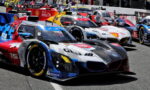 Lineup of colorful race cars in a pit lane, sponsor logos visible on the livery.