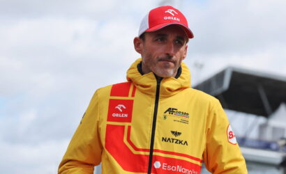 Male race car driver wearing a bright yellow jacket with red geometric patterns and sponsor logos, plus a red Orlen cap, standing outdoors near a racetrack.