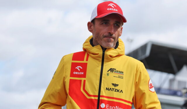 Male race car driver wearing a bright yellow jacket with red geometric patterns and sponsor logos, plus a red Orlen cap, standing outdoors near a racetrack.