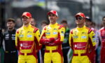 Three yellow-red race drivers stand with arms crossed in a pit lane, matching sponsor logos visible on their suits: Esa NanoTech and Michelin.