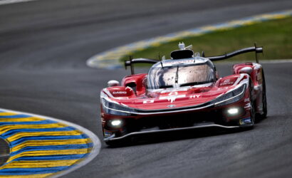 Red race car #8 speeds through a wet, curved track with blue-and-yellow curb on the inside. Headlights are on.