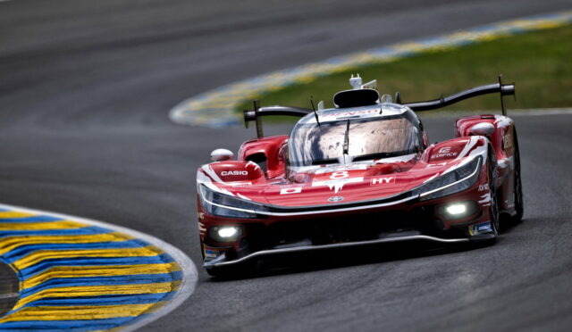Red race car #8 speeds through a wet, curved track with blue-and-yellow curb on the inside. Headlights are on.