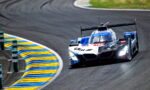Blue and white BMW race car rounding a blue-yellow curbed corner on a racetrack at speed (Prototype/Le Mans style).