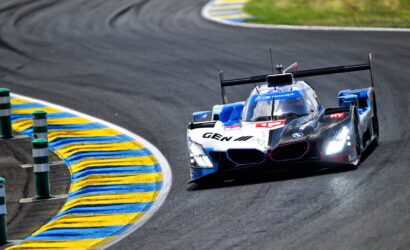 Blue and white BMW race car rounding a blue-yellow curbed corner on a racetrack at speed (Prototype/Le Mans style).