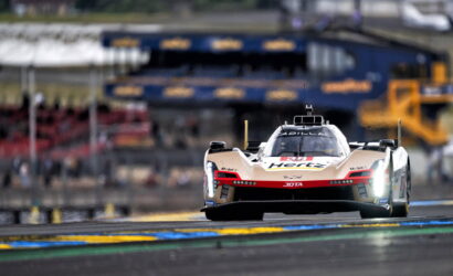 Low-angle front view of a white and red Cadillac race car speeding toward the camera on a race track, with blurred pit area in the background.