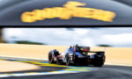 Blue Cadillac race car rounds a hairpin on a sunlit race track, with yellow/blue curbs and a blurred sponsor arch overhead—emphasizing speed and motion.