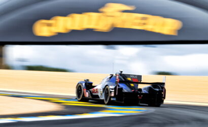 Blue Cadillac race car rounds a hairpin on a sunlit race track, with yellow/blue curbs and a blurred sponsor arch overhead—emphasizing speed and motion.