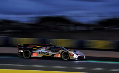 Blurry race car in Hertz livery speeding along a track at dusk, number 12 visible on the side, headlights on, with blurred barriers in the background.