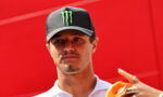 Man wearing a black cap with a green Monster logo and a white polo, against a bright red wall behind him.