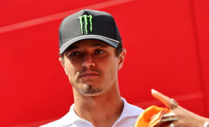 Man wearing a black cap with a green Monster logo and a white polo, against a bright red wall behind him.