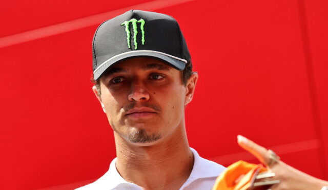 Man wearing a black cap with a green Monster logo and a white polo, against a bright red wall behind him.