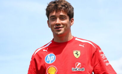 Young man in a red Ferrari shirt with sponsor logos, smiling against a blue sky.