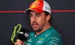 Man in a teal Aston Martin team shirt gives a press interview, speaking into a black FIA microphone.”] ,