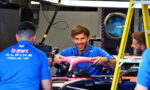 Team members in blue polo shirts work on a Formula 1 car in a garage; the smiling driver holds a pink front wing.
