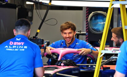 Team members in blue polo shirts work on a Formula 1 car in a garage; the smiling driver holds a pink front wing.