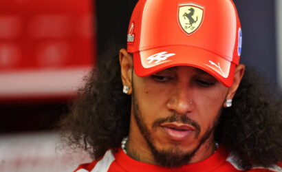 Man in a red Ferrari cap looking down, close-up portrait with earrings and beard.