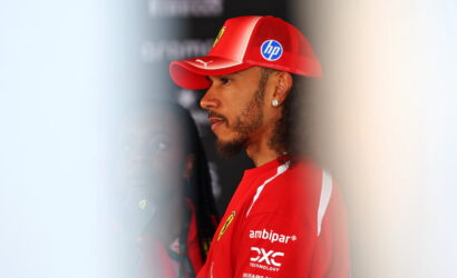 Side profile of a man in a red Ferrari team cap and shirt, facing left in a backstage area.