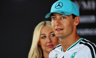 Man in a white Mercedes racing jersey and turquoise cap gazing to the right, with a blonde woman behind him at a media event.