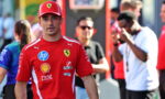 Man in a red Ferrari team shirt and cap walks toward the camera at a busy event, sponsor logos visible on his shirt.