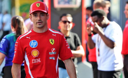 Man in a red Ferrari team shirt and cap walks toward the camera at a busy event, sponsor logos visible on his shirt.