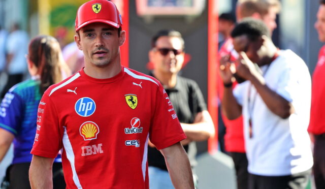 Man in a red Ferrari team shirt and cap walks toward the camera at a busy event, sponsor logos visible on his shirt.