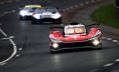 Red Formula-style race car leading a pack on a winding track with headlights on and blurred rivals behind.