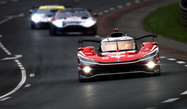 Red Formula-style race car leading a pack on a winding track with headlights on and blurred rivals behind.