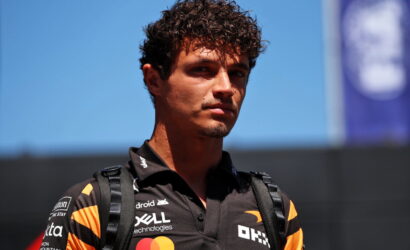 Young man with curly hair wearing a black polo shirt with sponsor logos and a backpack, standing outdoors against a blue sky.