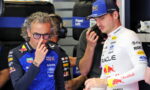 Two Formula 1 drivers in a garage; left in blue Red Bull Racing suit with glasses, right in white Oracle Racing suit.