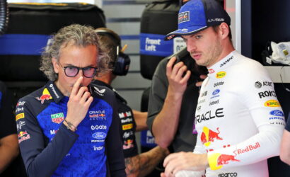 Two Formula 1 drivers in a garage; left in blue Red Bull Racing suit with glasses, right in white Oracle Racing suit.