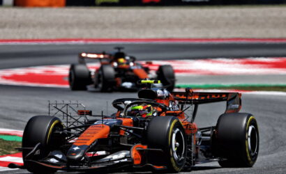 Orange and black Formula 1 car leading a race on a curved track with a second car behind it.