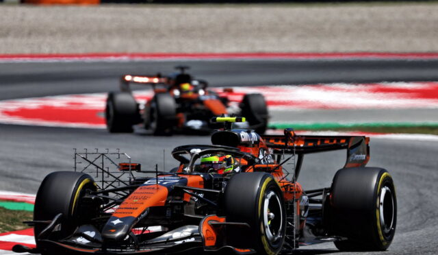 Orange and black Formula 1 car leading a race on a curved track with a second car behind it.