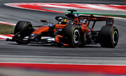 Orange Formula 1 car taking a tight corner on a race track, with motion blur of the curbs and asphalt behind it.