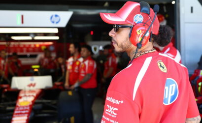 Side profile of a Ferrari pit crew member in a red cap and headset, standing in the pit lane with team members in the background, HP logo visible on the shirt.