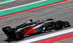 Red and silver Formula 1 car with Revolt branding speeds along a race track with green and red run-off lines in the background.