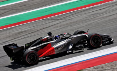 Red and silver Formula 1 car with Revolt branding speeds along a race track with green and red run-off lines in the background.