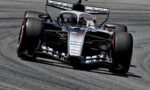 Silver and black Formula 1 car skids through a corner on a gray track, large rear wing and sponsor logos visible.
