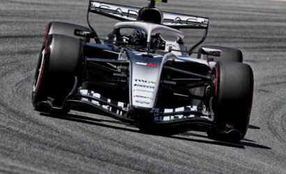 Silver and black Formula 1 car skids through a corner on a gray track, large rear wing and sponsor logos visible.