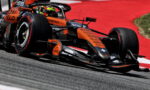 Orange Formula 1 race car leans into a corner on a red-tinted track surface, speed lines implied by motion blur