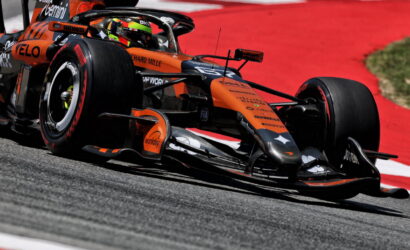 Orange Formula 1 race car leans into a corner on a red-tinted track surface, speed lines implied by motion blur