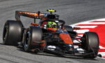 Orange and black Formula 1 race car rounding a left-hand turn on a gray track with a red curb in the background.