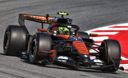 Orange and black Formula 1 race car rounding a left-hand turn on a gray track with a red curb in the background.